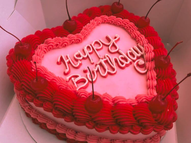 The image shows a pink heart-shaped birthday cake with red frosting and cherries on it